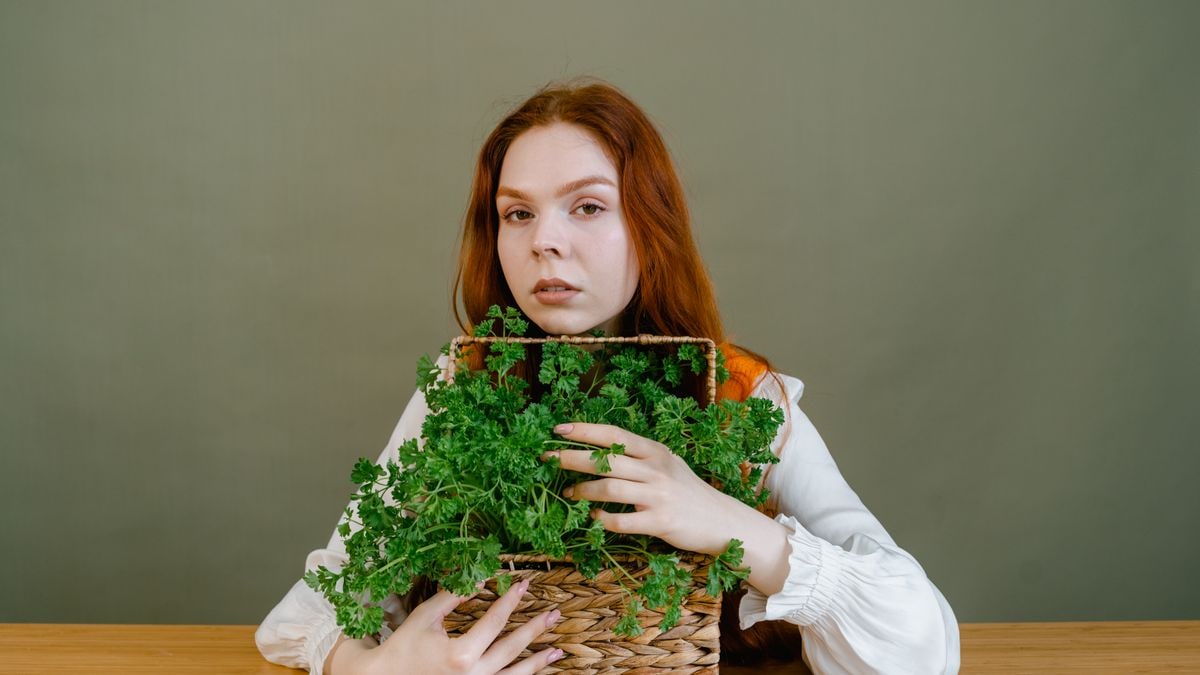 A red-headed woman holding a basket of fresh parsley against a gray background.
